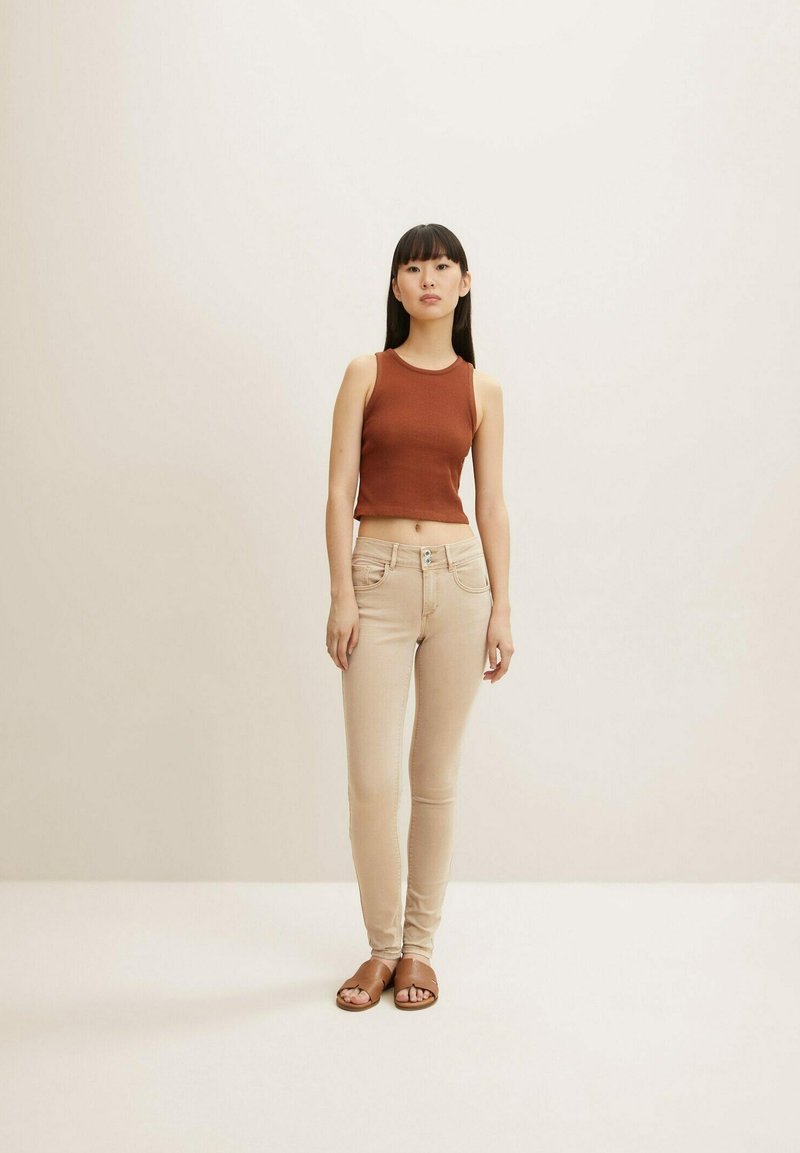 Rostfarbiges Tanktop kombiniert mit hellbeigen Skinny Jeans und braunen Riemchensandalen. Glattes Material und figurnahes Design, das die klare Silhouette betont.