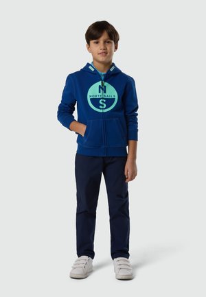 Ragazzo in piedi con una mano in tasca, indossa una felpa con zip blu North Sails, pantaloni blu scuro e sneaker bianche con chiusura a strappo su uno sfondo uniforme.
