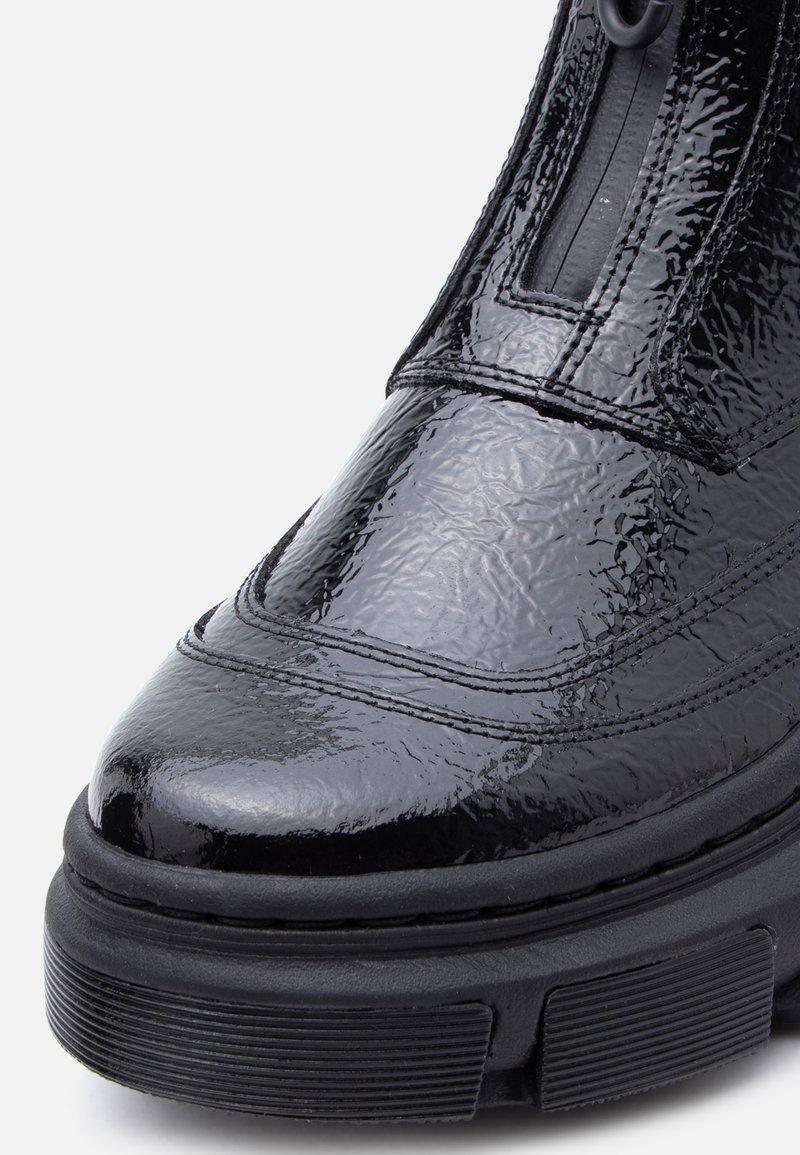 Close-up van een glanzende zwarte leren laars met gestructureerde details en dikke zwarte rubberen zool op een witte achtergrond.