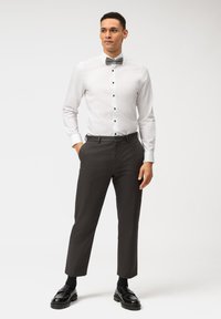 Camicia bianca con bottoni neri e papillon grigio, abbinata a pantaloni grigio scuro e mocassini neri, in posa in piedi.