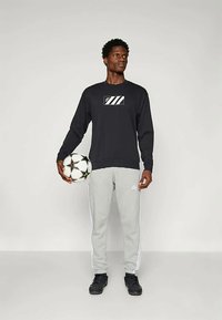 Maglietta nera a maniche lunghe con logo, pantaloni sportivi grigi con strisce bianche, sneakers nere, tiene in mano un pallone da calcio bianco e nero.