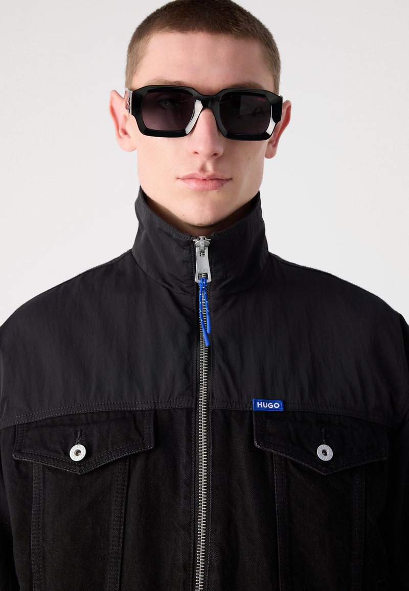 Czarna kurtka z wysokim kołnierzem, srebrnym zamkiem błyskawicznym oraz niebieskim logo. Posiada dwie kieszenie na piersi z srebrnymi guzikami. Czarne okulary przeciwsłoneczne z grubymi oprawkami.