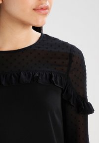 Sort bluse med et gennemsigtigt prikket stof og en rynket halsudskæring. Ærmerne har et delikat, flæset design og en glat tekstur.