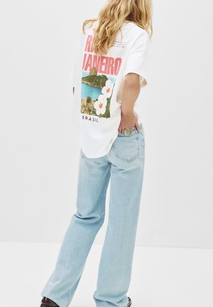 T-shirt graphique blanc avec un imprimé d'un paysage côtier et de fleurs, associé à un jean large bleu clair. Accent en argent avec une bague circulaire.