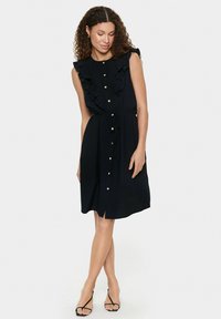 Abito nero con colletto arricciato, design senza maniche, bottoni frontali e vita con lacci. Tessuto liscio con orlo lungo fino al ginocchio e spacco laterale.