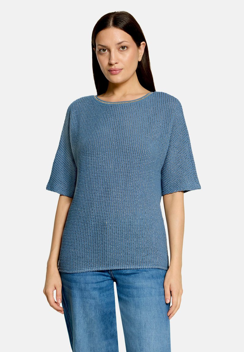 Femme aux longs cheveux foncés portant un haut bleu texturé à manches courtes et un jean bleu, debout devant un fond clair uni.