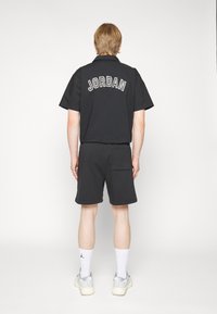 Svart kortärmad skjorta med "JORDAN" broderat på ryggen, parat med svarta shorts och vita strumpor, kombinerat med grå sneakers.