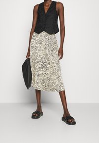 Sort tekstureret ærmeløs vest, flydende nederdel med sort og creme abstrakt mønster, sorte flade sandaler og en sort håndtaske.