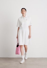 Balta polo suknelė su trumpomis rankovėmis ir elastinga juosmia. Modelis laiko rožinį maišą, derinama su baltomis sneaker'iais ir kulkšninių kojinių. Lygus audinys.
