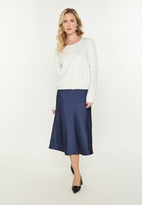 Witte gebreide cardigan met knoopsluiting en lange mouwen, gecombineerd met een marineblauwe satijnen midi-rok. Zwarte puntige hakken maken de look af.
