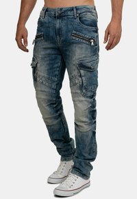 Pantaloni cargo in denim blu con numerose tasche con zip, una texture sbiadita e cuciture rinforzate. Indossati con sneakers bianche.