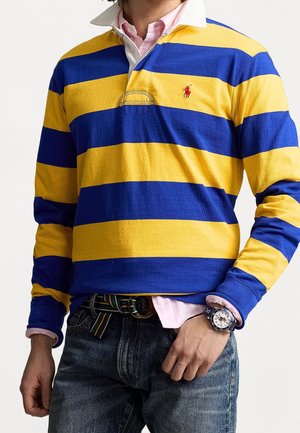 Niebiesko-żółta, paskowana koszulka polo z długim rękawem i białym kołnierzykiem. Dżinsy z kolorowym paskiem. Srebrny zegarek na nadgarstku.