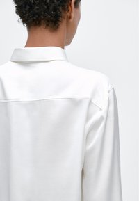 Camicia bianca con collo a camicia, tessuto liscio e vestibilità rilassata. Presenta una cucitura centrale sulla schiena e spalle calate.