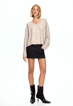 Beige, kurz geschnittenes Sweatshirt aus weichem Material, kombiniert mit einem dunklen Jeans-Minirock. Schwarze, spitze Stiefeletten mit Reißverschlussdetails.