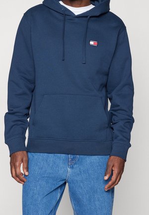 Mężczyzna w granatowej bluzie Tommy Jeans z kapturem i jasnoniebieskich dżinsach stoi na jednolitym jasnym tle.