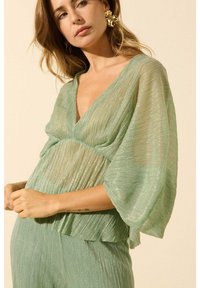 Haut plissé vert transparent avec décolleté en V et manches larges ; tissu texturé avec des reflets ; ourlet superposé ; porté avec un bas assorti.
