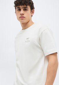 Νεαρός άνδρας με σγουρά μαλλιά που φοράει ένα απλό λευκό κοντομάνικο μπλουζάκι με μικρό λογότυπο "Marc O’Polo" στο στήθος.