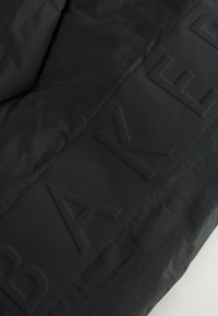 Tissu noir matelassé avec une texture lisse, présentant un logo "BAKER" en relief dans une bande solide, soulignant un design minimaliste.