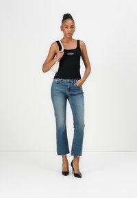 Schwarzes Tanktop mit Logo, blaue hochgeschnittene Jeans mit leichtem Schlag und schwarze spitze High Heels. Modell hält eine kleine weiße Handtasche.