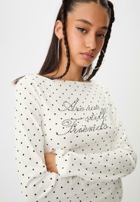 Biela košeľa s dlhými rukávmi zdobená čiernymi puntíkmi, s textovým dizajnom z rhinestones na prednej strane, ktorý znie "Sme ešte priatelia?"