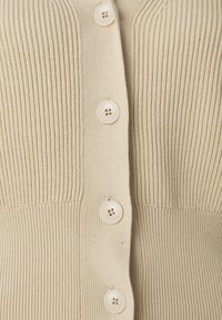 Beige ribbestrikket cardigan med fem runde, lyse knapper foran, med en myk tekstur og en kroppsnær design.