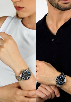 Due orologi in acciaio inossidabile argentato con quadranti neri e accenti blu, indossati ai polsi di una persona con una maglietta bianca e di un'altra con una polo nera.