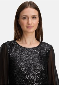 Jonge vrouw met steil bruin haar, gekleed in een zwarte top met pailletten en doorzichtige zwarte lange mouwen, met een neutrale uitdrukking, op een witte achtergrond.
