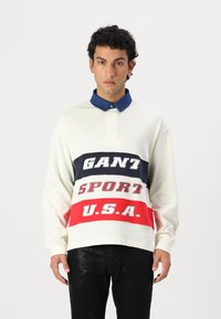 Langermet polo skjorte i krem med blå krage. Har iøynefallende grafikk i marineblå, rød og grå med teksten "GANT SPORT U.S.A." foran.