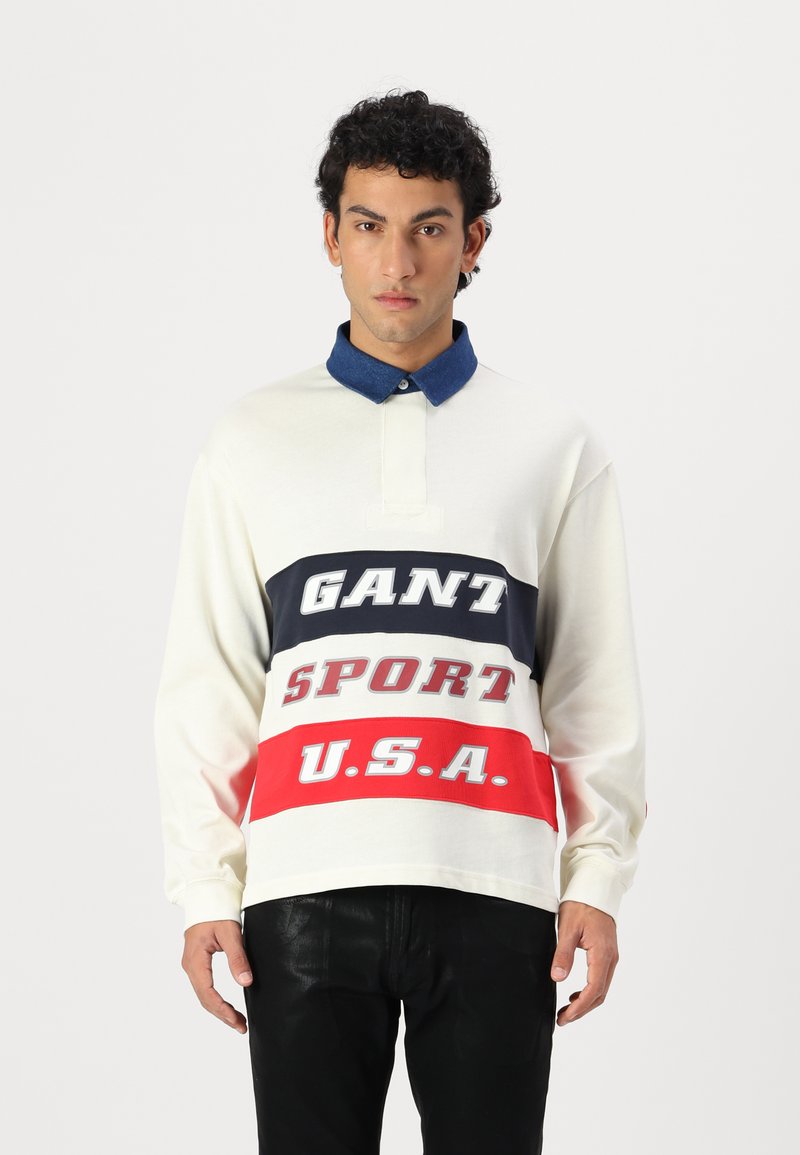 Langermet polo skjorte i krem med blå krage. Har iøynefallende grafikk i marineblå, rød og grå med teksten "GANT SPORT U.S.A." foran.