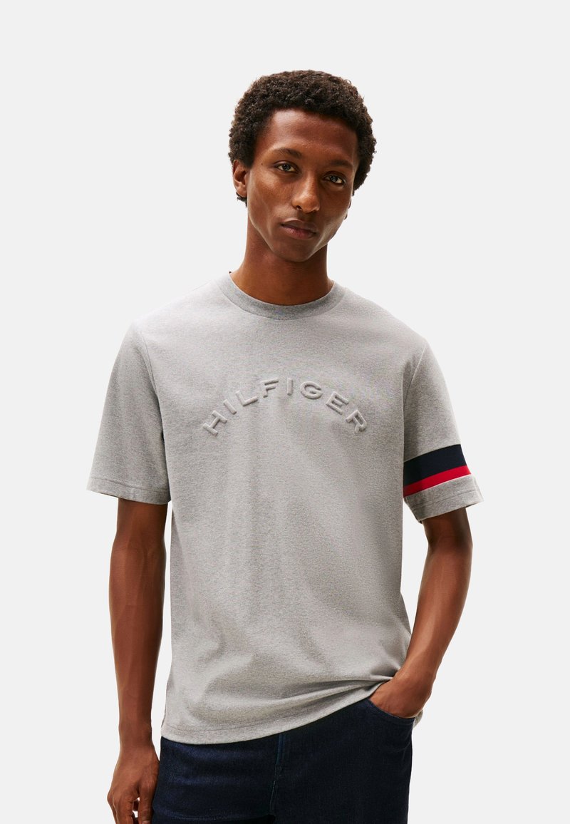 Camiseta de algodón gris claro con el logo bordado "HILFIGER", mangas cortas y un acento con franjas azul marino, rojo y blanco en los puños.