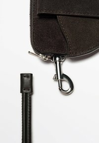 Portefeuille en daim marron avec une fermeture éclair et un fermoir en métal. Dispose d'une sangle en cuir lisse sur la gauche, mettant en avant un design épuré et un savoir-faire de qualité.