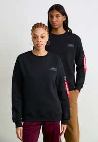 Zwarte sweatshirt met een ronde hals, gestructureerde stof, zijzak met rits en rode label, en logo patch op de voorkant. Gemoodelleerd door twee personen.