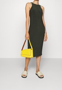 Žena v bezramenných tmavozelených midi šatech, která drží žlutou kabelku s hnědým popruhem, na nohou má béžové sandály na klínku, na jednobarevném pozadí.