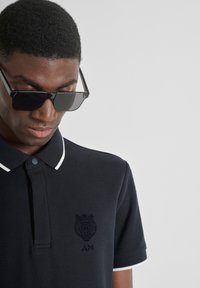 Crna polo majica s bijelim ovratnikom i obrubom. Ima izvezeni logo lava i inicijale "AM" na prsima. Glatka tekstura tkanine.