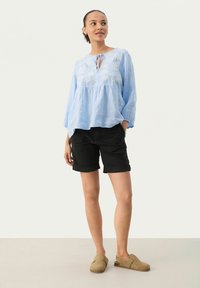 Lyseblå langærmet bluse med broderet blomsterdesign og snørering ved halsen, parret med sorte shorts og beige slip-on sko.