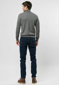Pull à col roulé gris, design ajusté, poignets côtelés, porté avec un jean bleu foncé, poches arrière visibles, et des bottes à lacets marron.