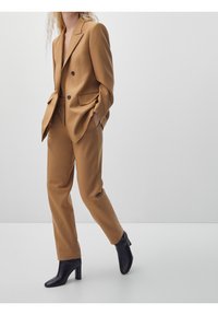 En person med vågigt hår bär en skräddarsydd brun kostym och svarta klackstövlar, poserande mot en minimalisk ljus bakgrund.