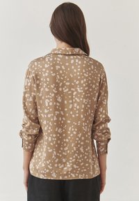 Bruine-shirt met onregelmatige witte vlekken, lange mouwen en een kraag. De stof lijkt glad met een lichte glans.
