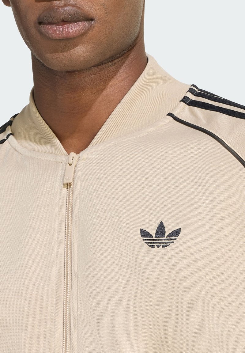 Beige zip-up jacket van een textuurstof, met zwarte strepen op de schouders en een klein zwart logo op de borst.