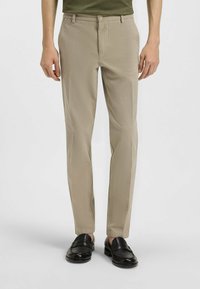 Beige op maat gemaakte broek met een gladde textuur, rechte pasvorm en een subtiel patroon. Voorzien van een ritssluiting en knoopsluiting aan de voorkant, gecombineerd met zwart leren instappers.