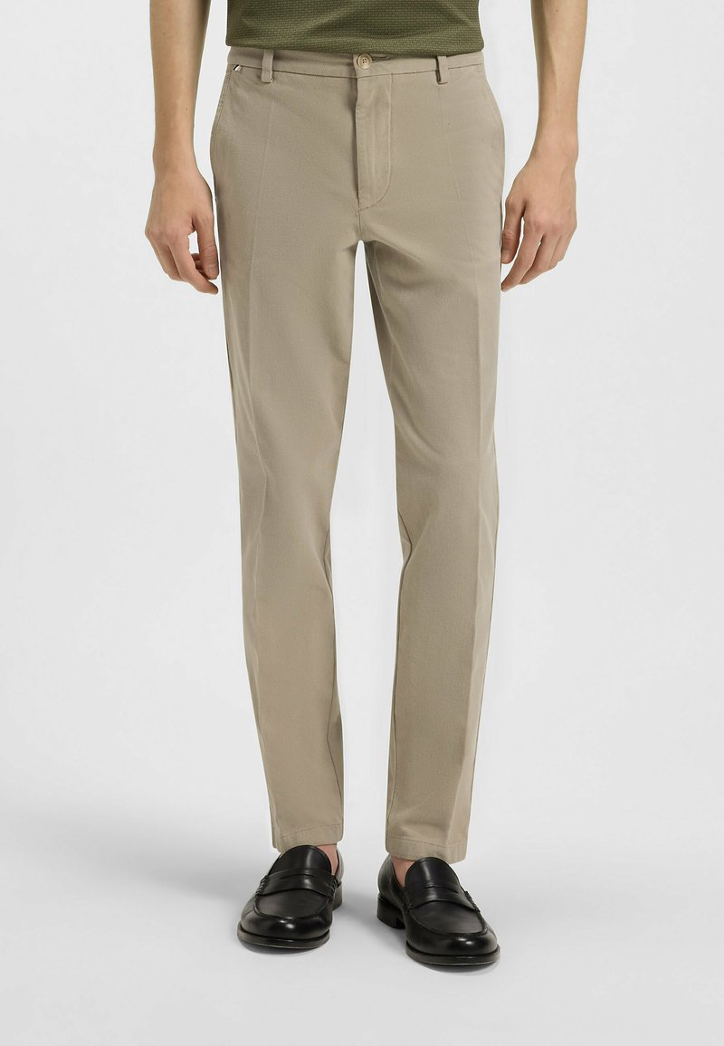 Beige op maat gemaakte broek met een gladde textuur, rechte pasvorm en een subtiel patroon. Voorzien van een ritssluiting en knoopsluiting aan de voorkant, gecombineerd met zwart leren instappers.