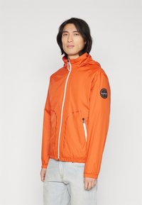 Uomo che indossa una giacca a zip arancione brillante con cappuccio e jeans azzurri chiari, in piedi contro uno sfondo chiaro e uniforme.