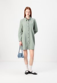 Abito button-up verde chiaro con maniche lunghe, caratterizzato da uno yoke curvo e bottoni neri. Abbinato a una borsa blu e ballerine nere.