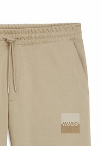 Beiga mjukisbyxor med elastisk midja och dragsko. Har sidofickor och en fyrkantig logotyp med ljusbrun färg och texten "HUGO".