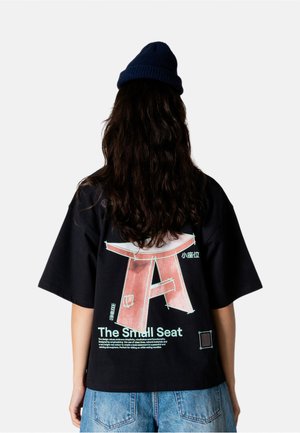 Person med langt mørkt hår iført en marineblå hue, sort t-shirt med en stor lyserød sædeillustration og tekst, samt blå denimshorts, set bagfra.