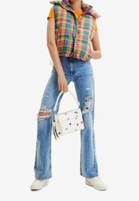 Bunte karierte Steppweste mit hohem Kragen, kombiniert mit ausgewaschenen blauen Jeans. Weiße Tasche mit floraler Stickerei, akzentuiert mit mehrfarbigen Quasten.