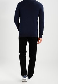 Mann trägt einen marineblauen Pullover, schwarze Hosen und weiße Sneakers und geht vor einem schlichten weißen Hintergrund weg.