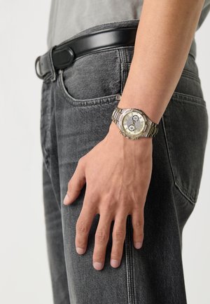 Sølv- og gullmetallisk klokke med rund urskive, svart skive og tre underdials. Båret på et håndledd, kombinert med mørkegrå jeans.
