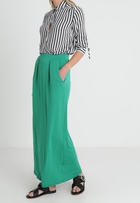 Pruhovaná černobílá blůza s límečkem v kombinaci s dlouhou, splývavou zelenou sukní s předními skladami a bočními kapsami. Černé sandály.