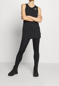 Czarny bezrękawnik sportowy z logo, zestawiony z czarnymi legginsami z siateczkowymi panelami oraz czarnymi, wytrzymałymi butami. Prosty, funkcjonalny design.